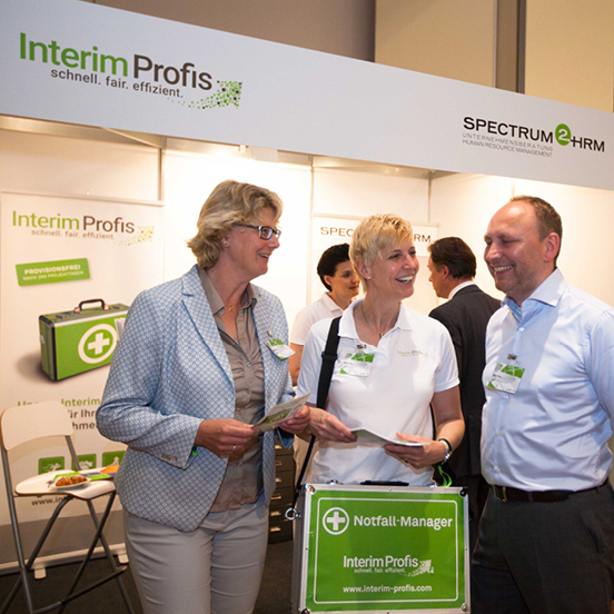 Annette Elias und ein Interim Manager und eine Interim Managerin stehen an einem Messestand der Interim Profis 