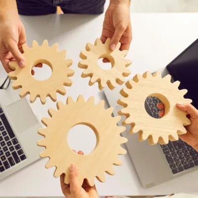 Zwei Personen stehen an einem Schreibtisch und halten hölzerne Zahnräder in den Händen, daneben liegen geöffnete Laptops.